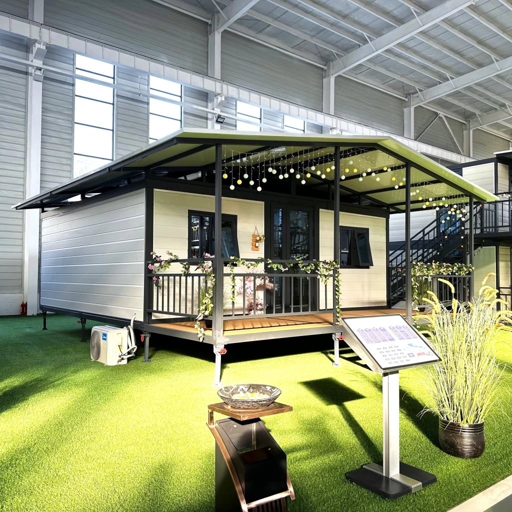 IMG_6126 Expandable home with a light exterior, dark framing, veranda and pitched roof, displayed inside a factory.